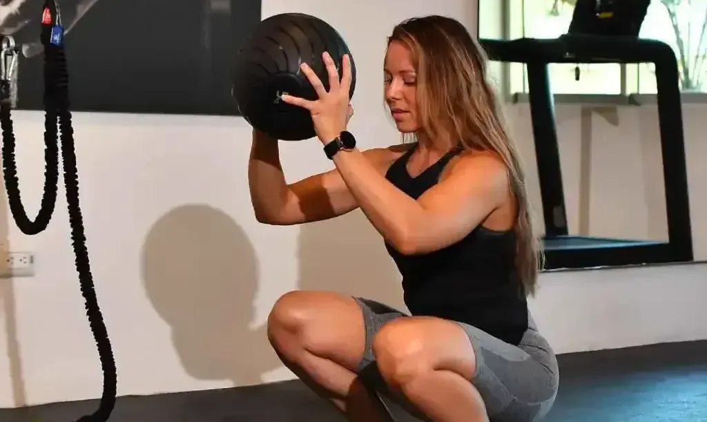 Woman in athletic wear squats, holding a medicine ball in a gym.