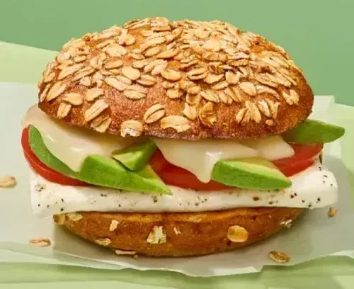 Oatmeal roll sandwich with avocado, tomato, cheese, and egg on a light green surface.