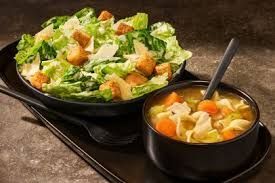 Caesar salad and chicken noodle soup in black bowls on a dark tray.