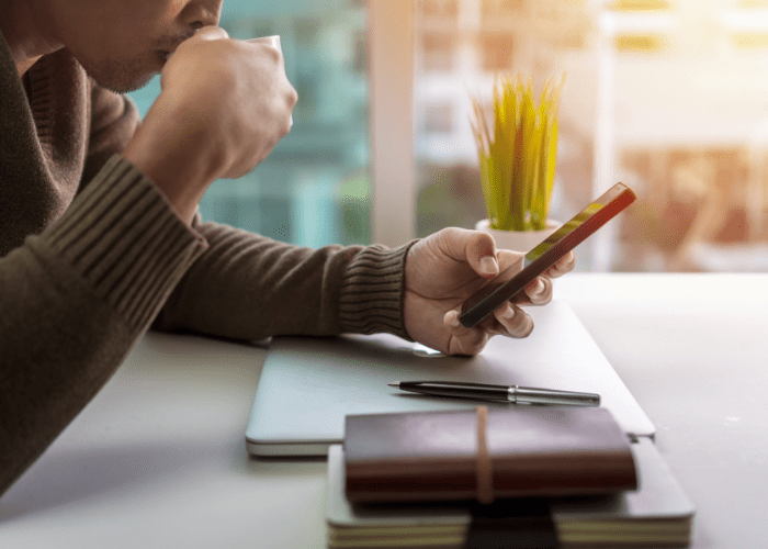 A man is sitting at a table drinking coffee and looking at his cell phone.