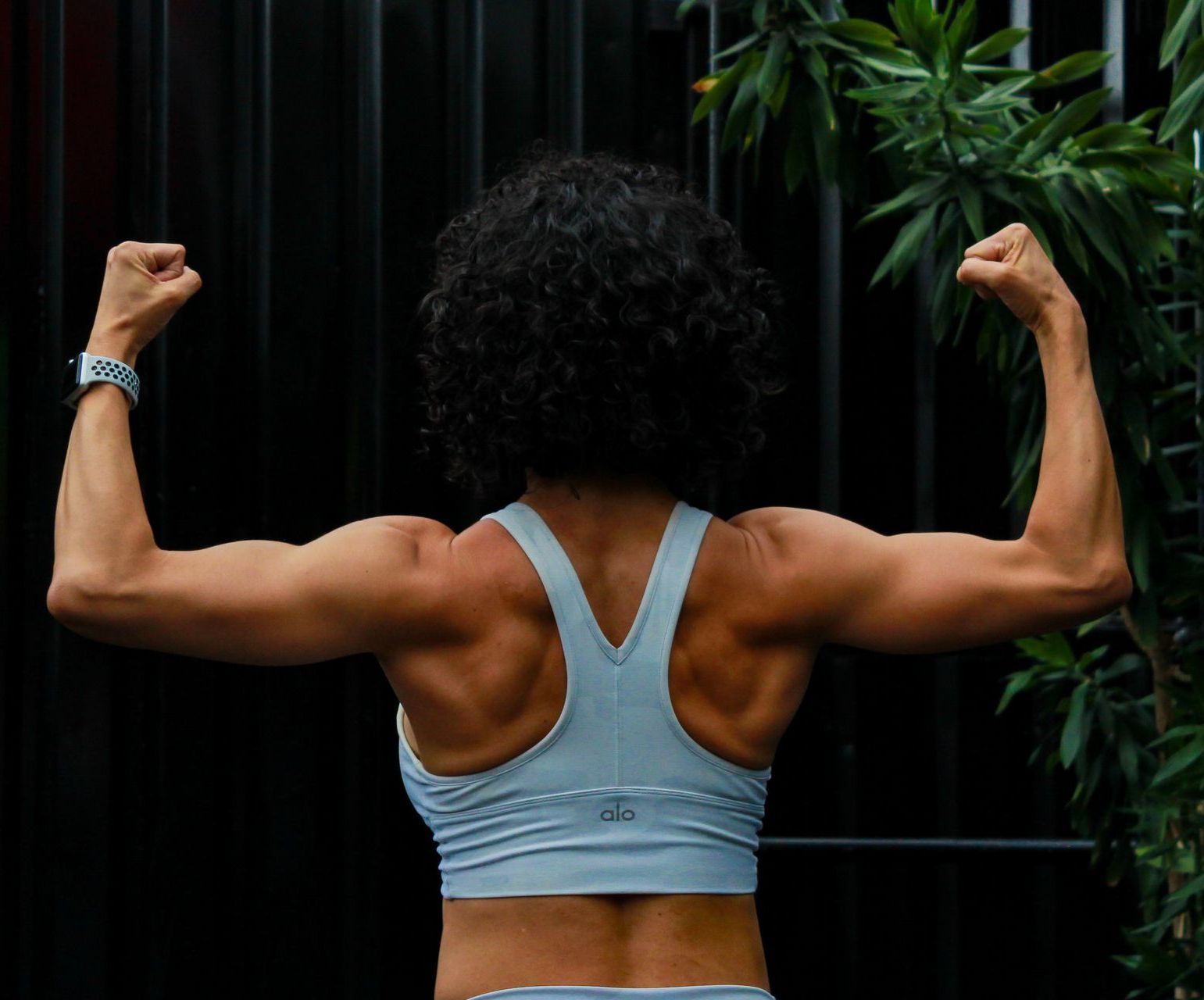 A woman is flexing her muscles in front of a black wall. Strength Training and Nutritional Coaching