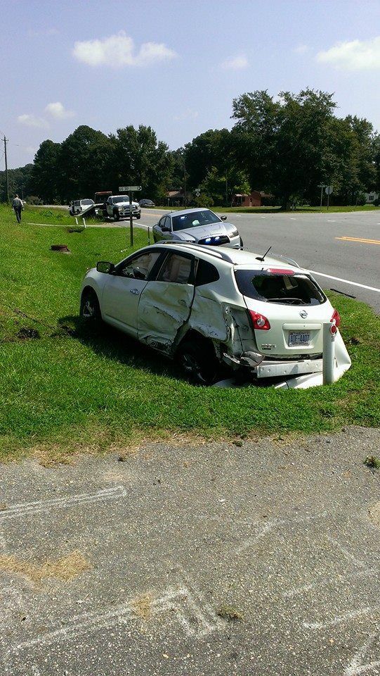 White car, damaged from an accident, sits in grass beside a road; police car and other vehicles visible in the background.
