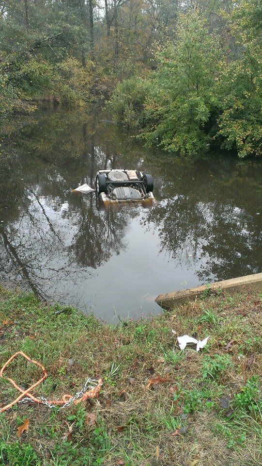 Car submerged in water, upside down. Green trees and foliage surround the water. Cloudy day.