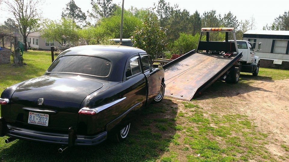Black classic car being towed onto a flatbed tow truck in a grassy yard.