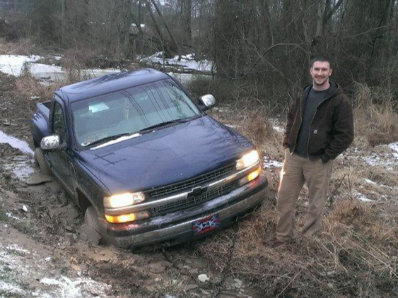 Man standing beside a blue pickup truck in a muddy, wooded area in rocky mount nc