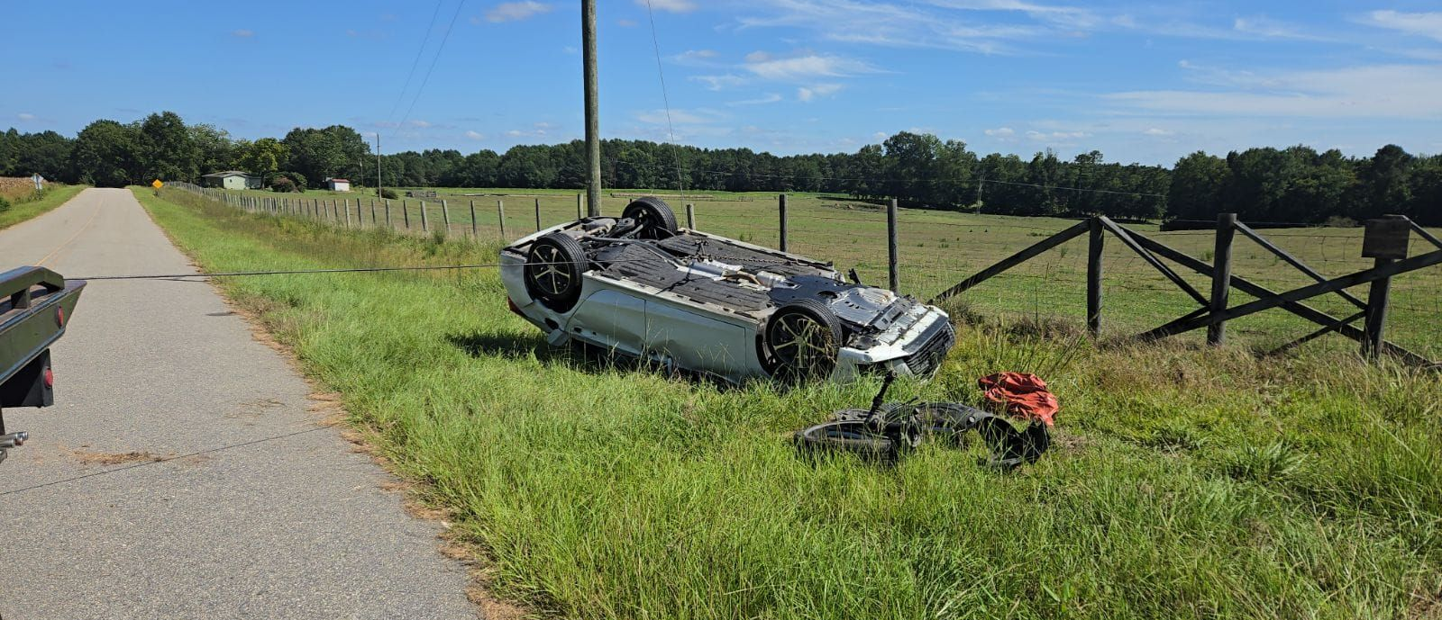 Car overturned in grassy ditch next to road, fence and telephone pole in background, sunny day in Rocky Mount, NC.