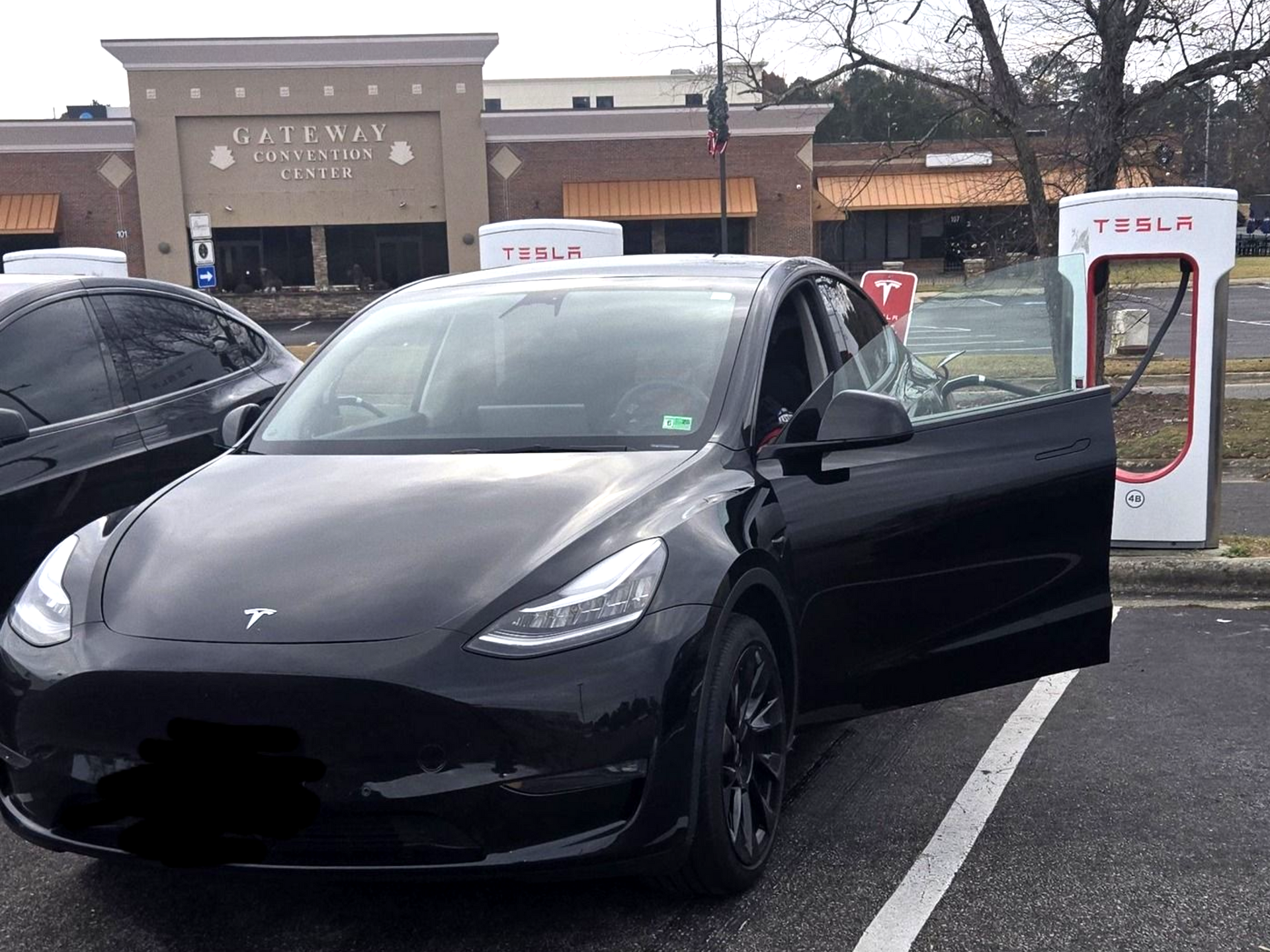 Black Tesla Model Y charging at a Supercharger station, door open, in front of a shopping center.