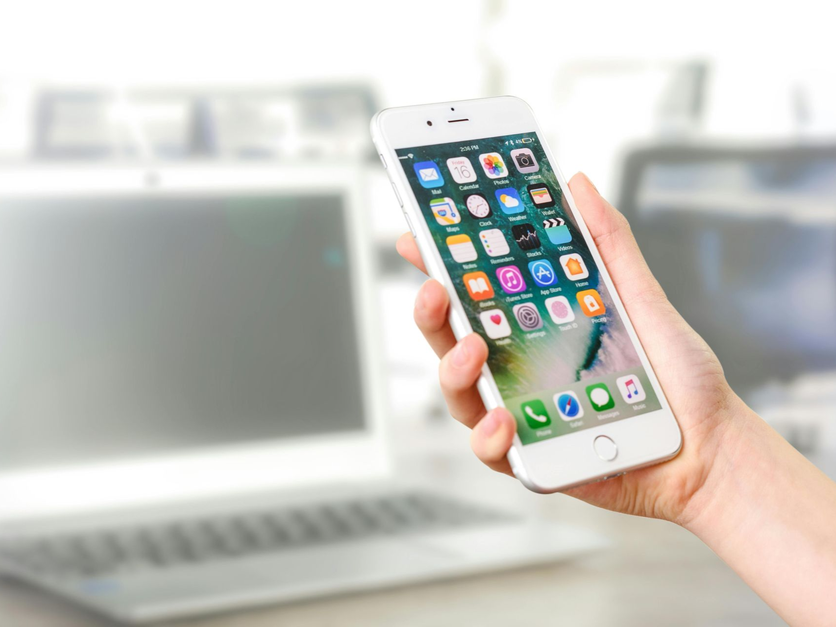 Hand holding a white smartphone displaying apps, in an office setting with a laptop in the background.