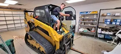 A person in a yellow skid steer in a garage. They are reaching inside the cab with the door open.