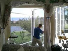 Man installing window screen in a room with lake view. Beige curtains, blue shirt.