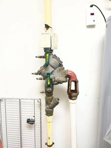 Industrial valve assembly with pipes and a brass valve on a white wall, with a gauge and labels.