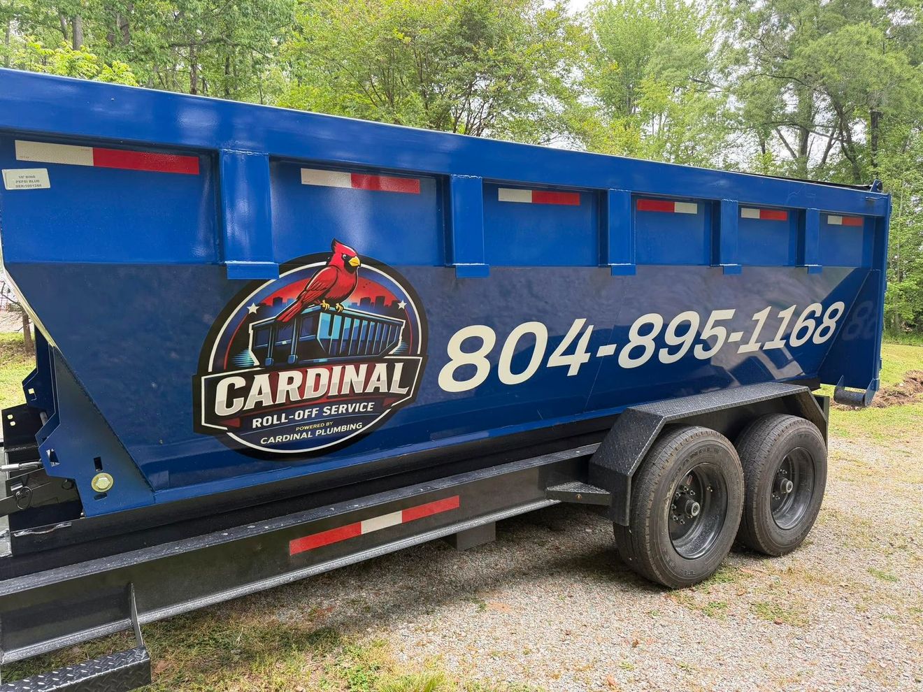 Blue Cardinal dump trailer with “804-895-1168” parked on gravel by trees