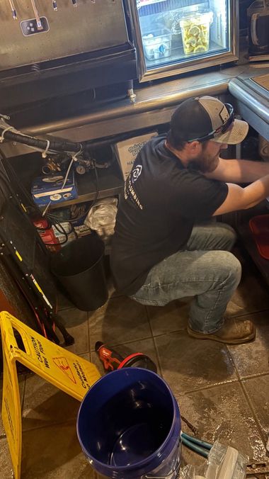 Worker crouches beside machinery in a dim workshop, with a blue bucket and tools in the foreground.