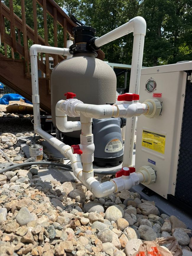 Pool equipment with white pipes, red valves, and a gray filter beside a house wall and rocks.