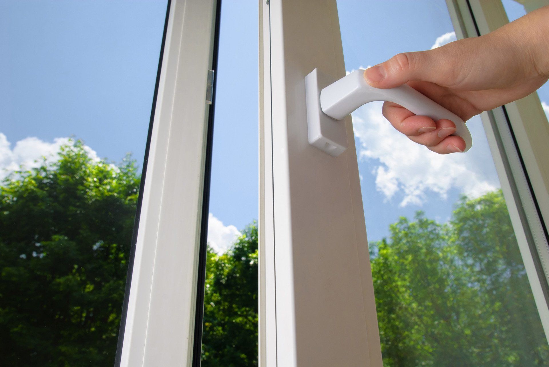 Hand opening a white window handle with a clear blue sky and green trees visible outside