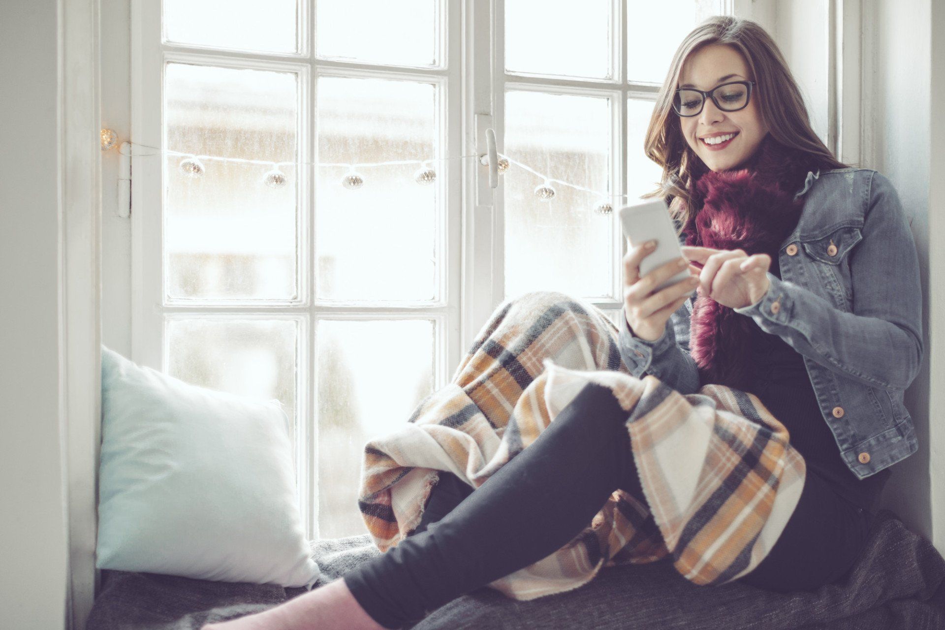 Woman wrapped in a blanket sitting by a window and smiling while using a smartphone