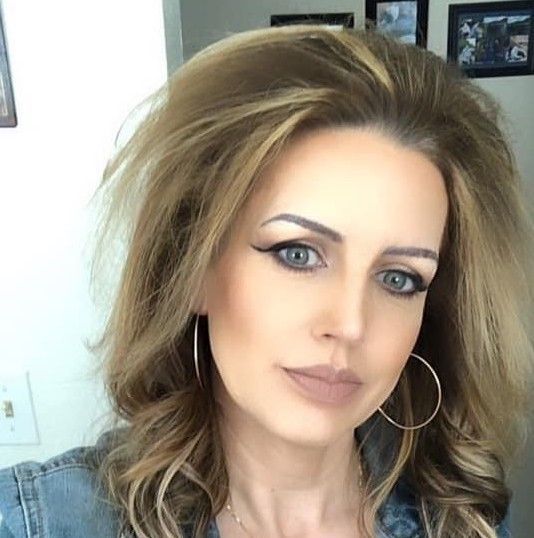 Close-up portrait of a woman with voluminous blonde hair, hoop earrings, and makeup, facing the camera indoors