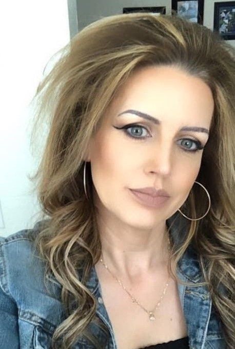 Portrait of a woman with voluminous blonde hair, hoop earrings, and a denim jacket, looking at the camera