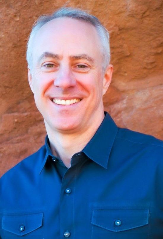 Smiling man in a navy shirt standing against a reddish rock wall.