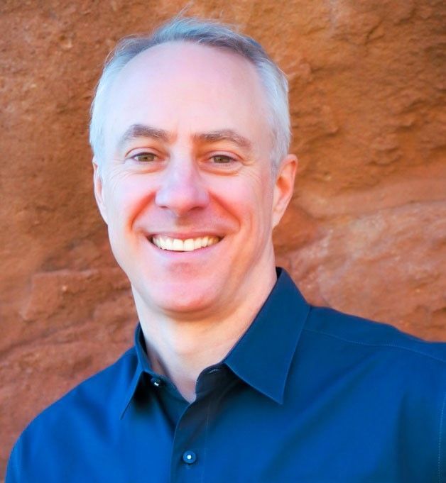 Smiling person in a dark blue shirt standing before a reddish rock wall