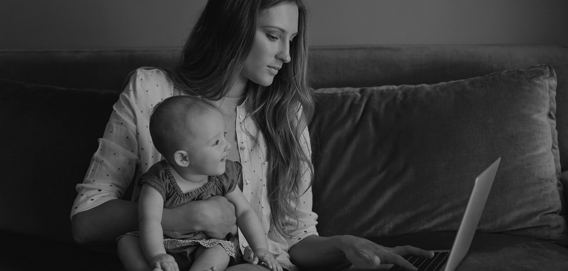Woman sitting on a couch with a baby, looking at a laptop in a dim living room