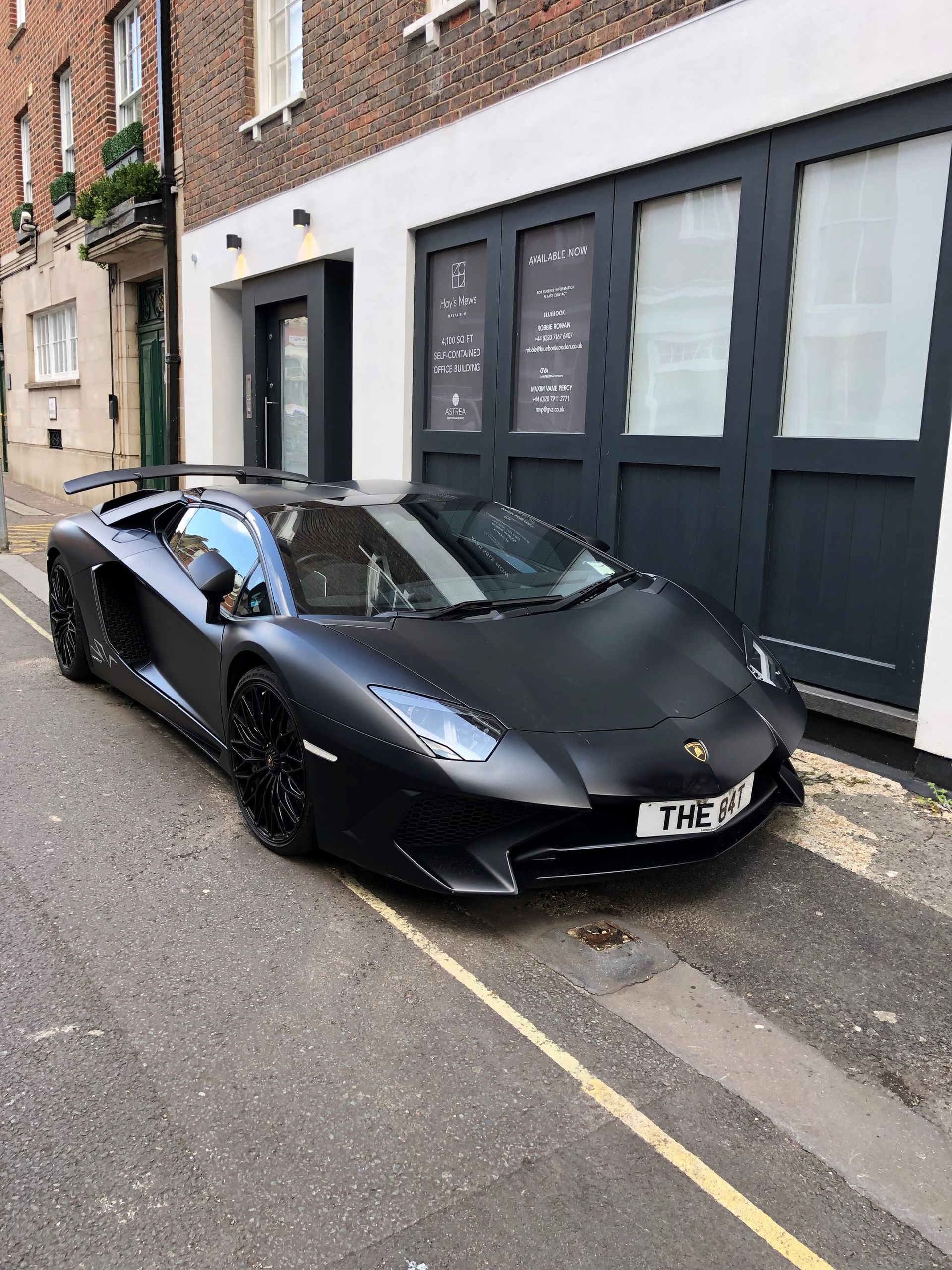 Black Lamborghini parked on a narrow street beside a brick building with dark storefront windows.