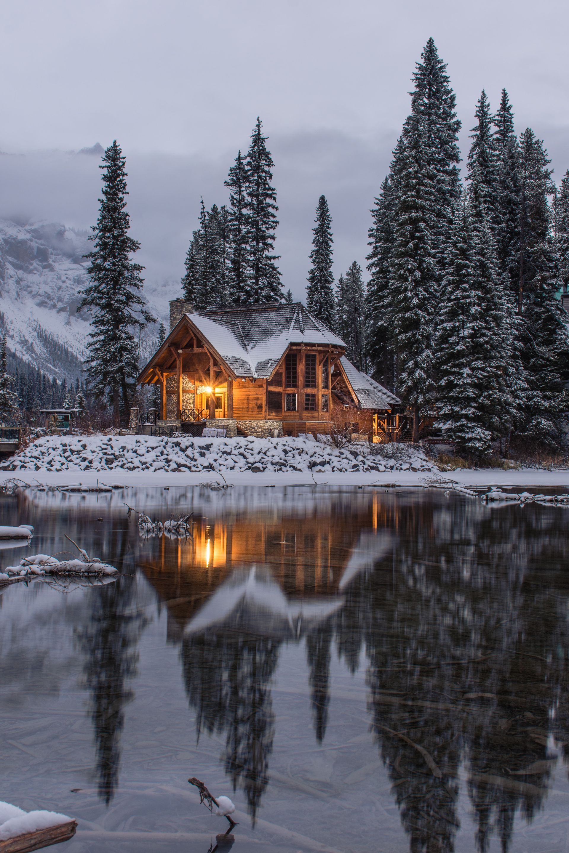 Snowy lakeside cabin glowing at dusk, reflected in calm water beside tall pine trees.