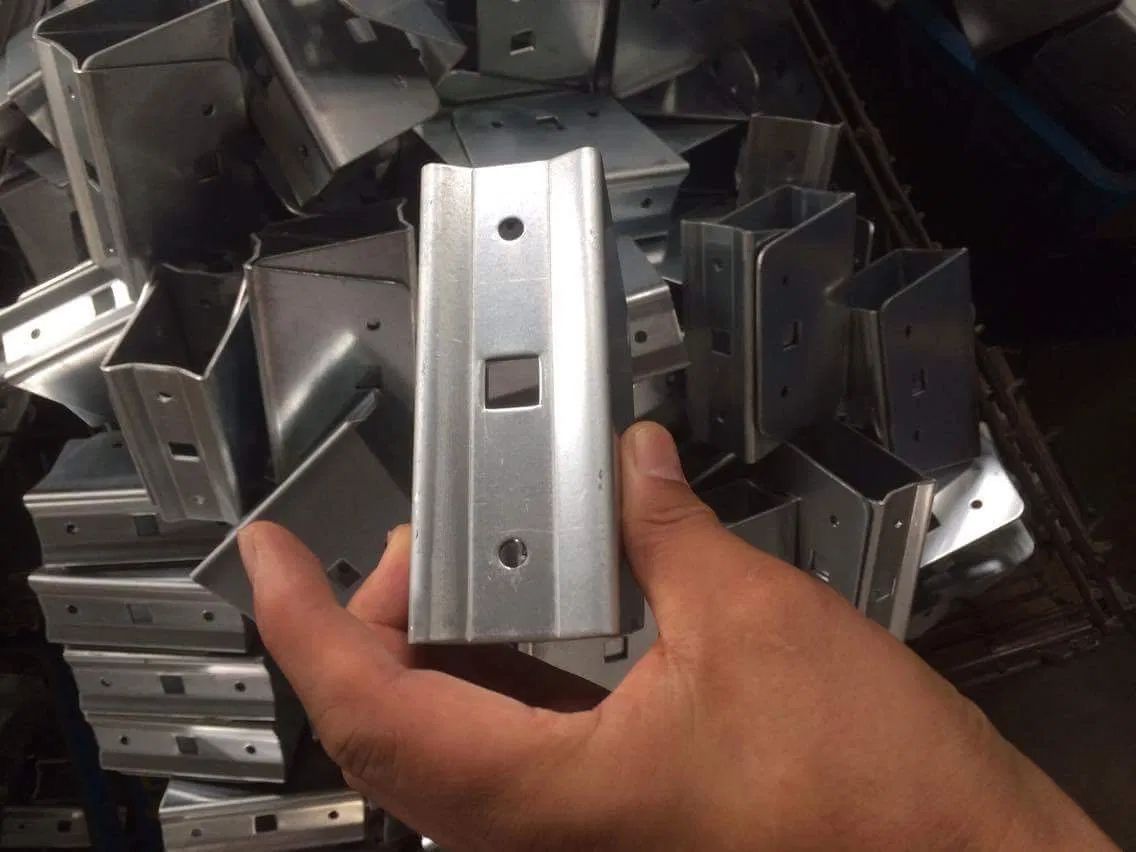 A hand holding a metallic, U-shaped bracket with circular and square holes, against a background of similar parts.