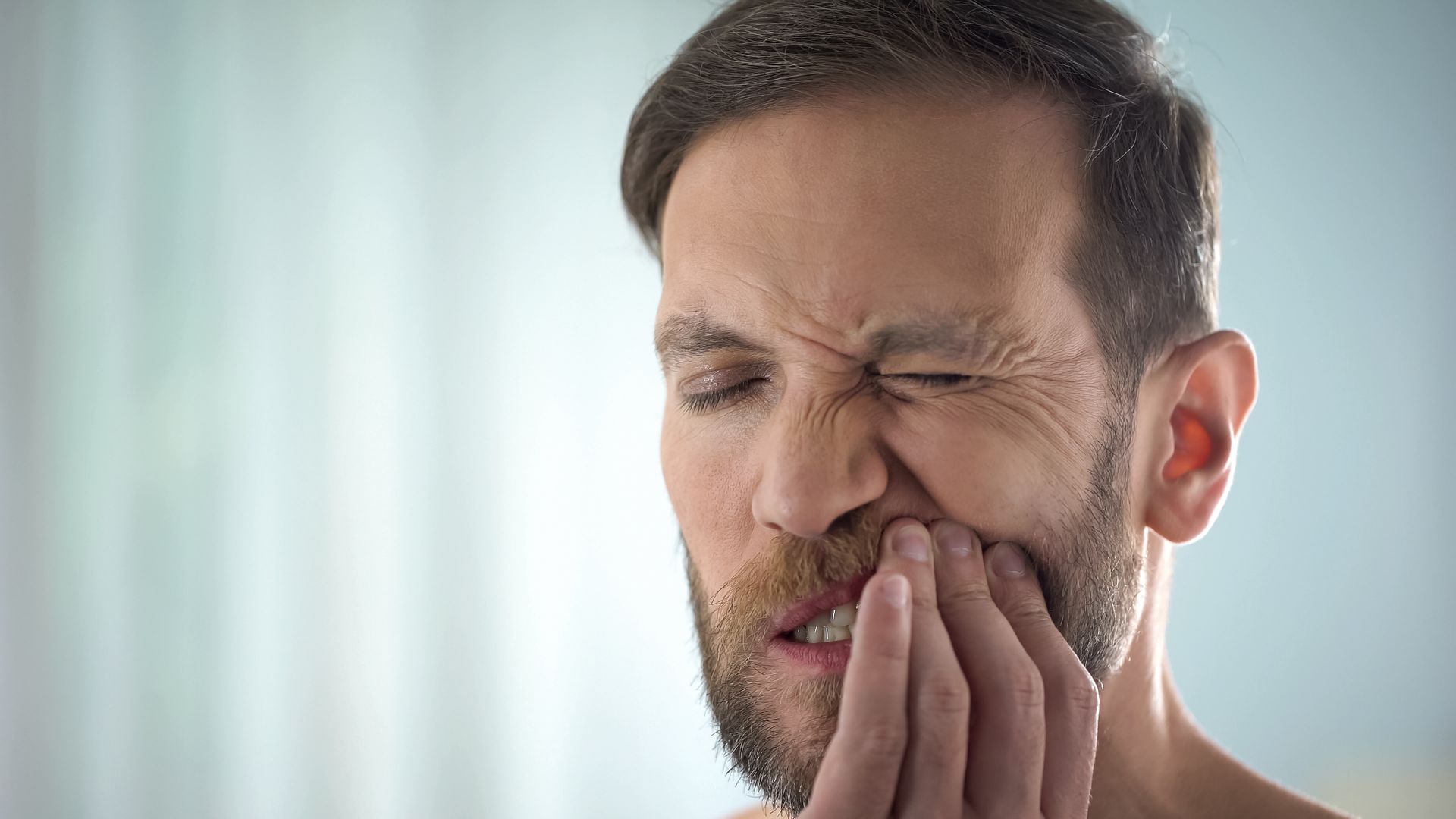 Man holding his cheek with eyes closed, grimacing in pain against a bright background