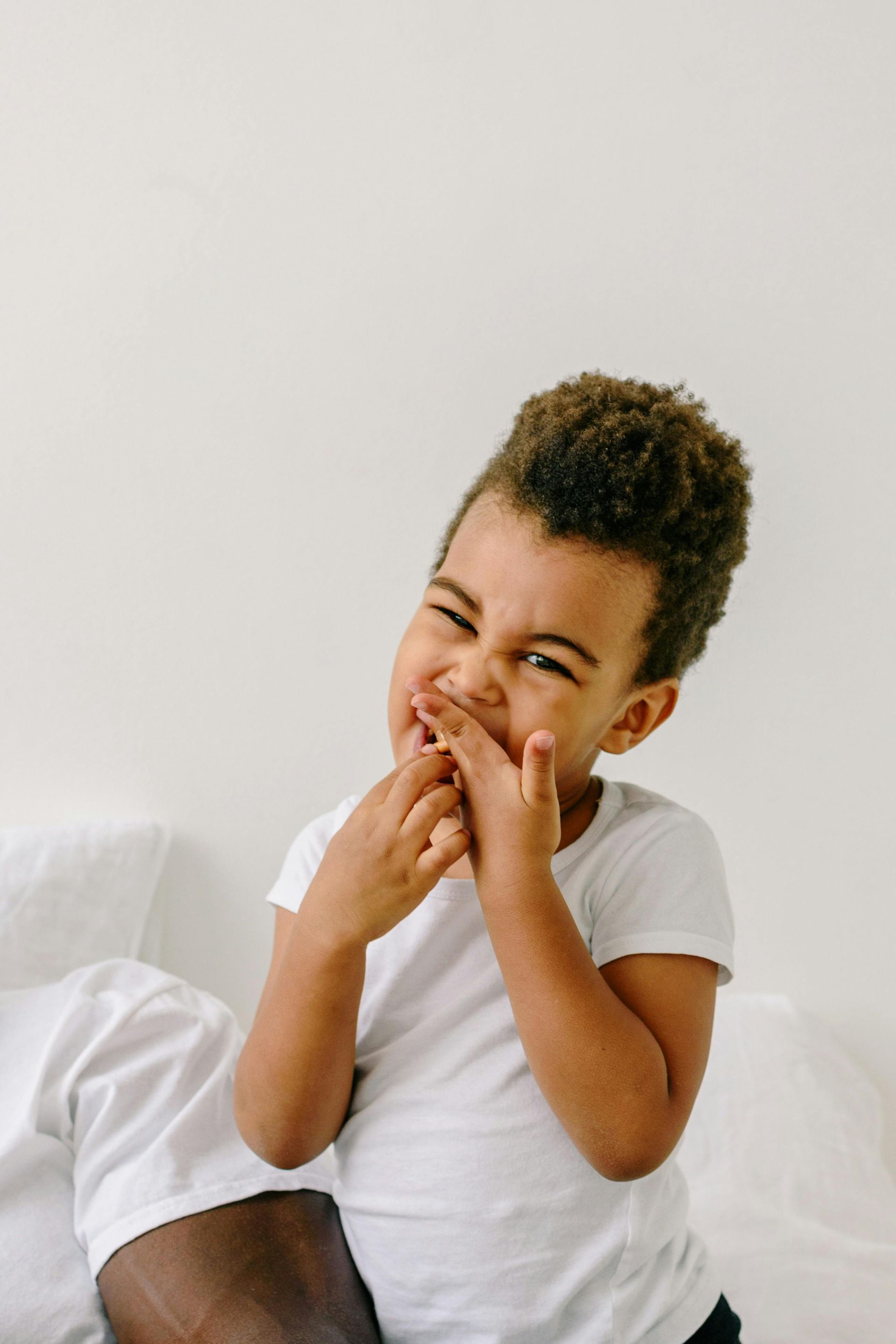Child laughing on a white bed, wearing a white shirt and covering their mouth with both hands.