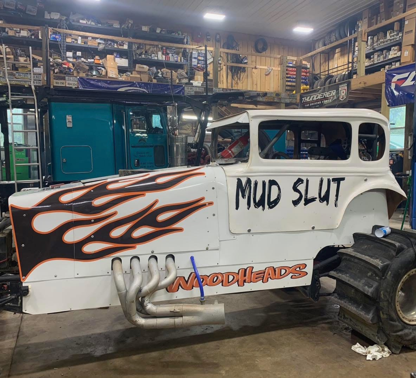 White mud truck with flame graphics and “MUD SLUT” lettering in a garage/workshop.