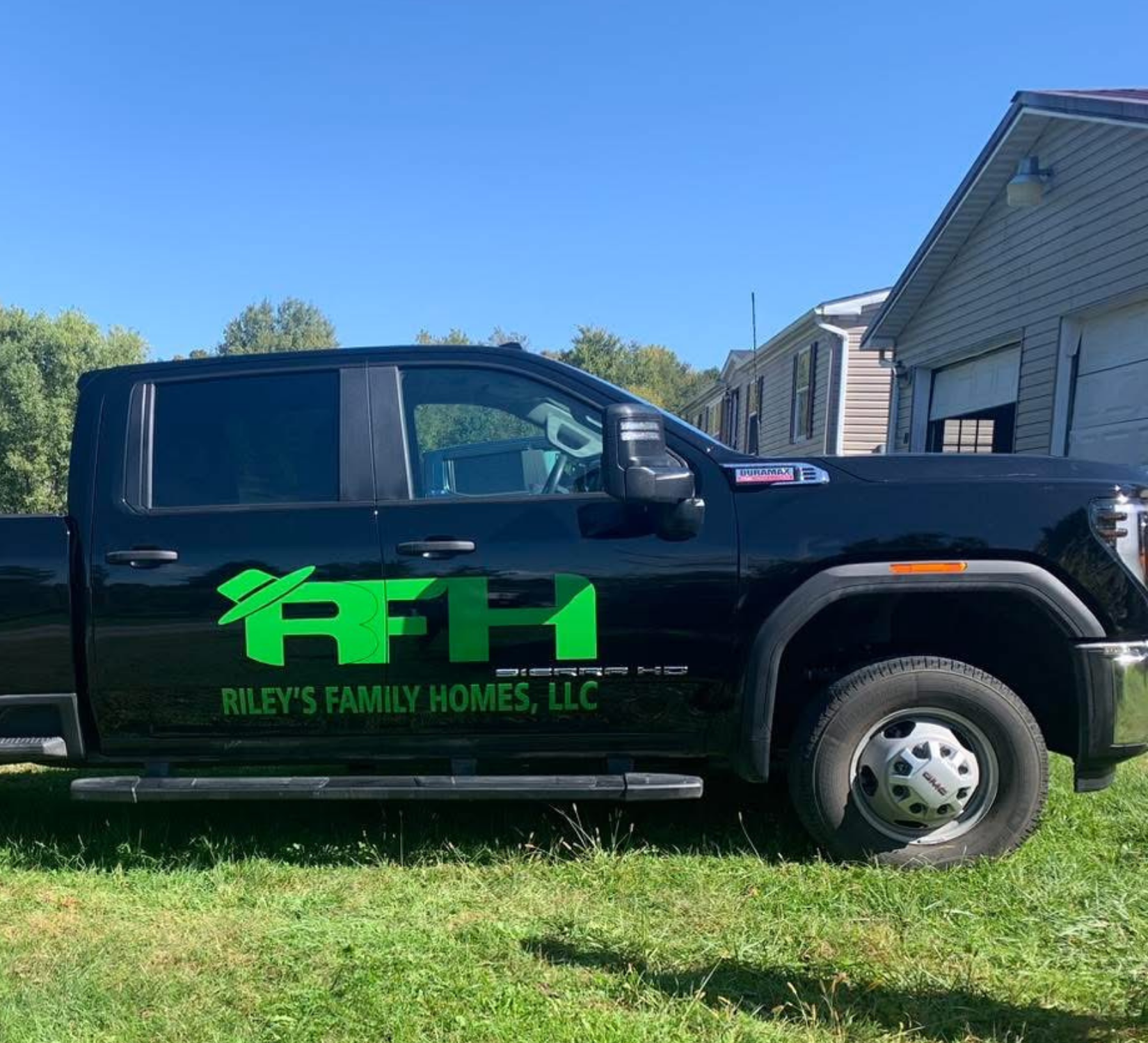 Black work truck with green “FTM” logo and “Riley’s Family Homes, LLC” parked on grass by a house