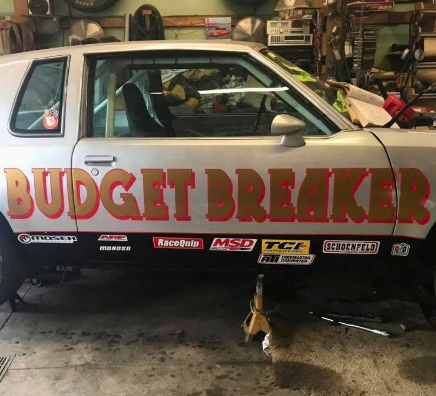Vintage race car in a garage with bold red “BUDGET BREAKER” lettering on the door.