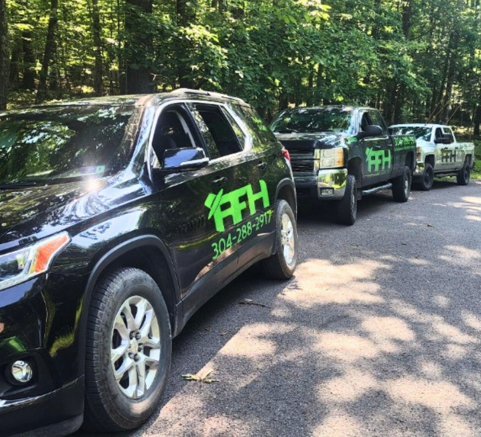 Three black off-road SUVs with green logos parked in a wooded roadside line