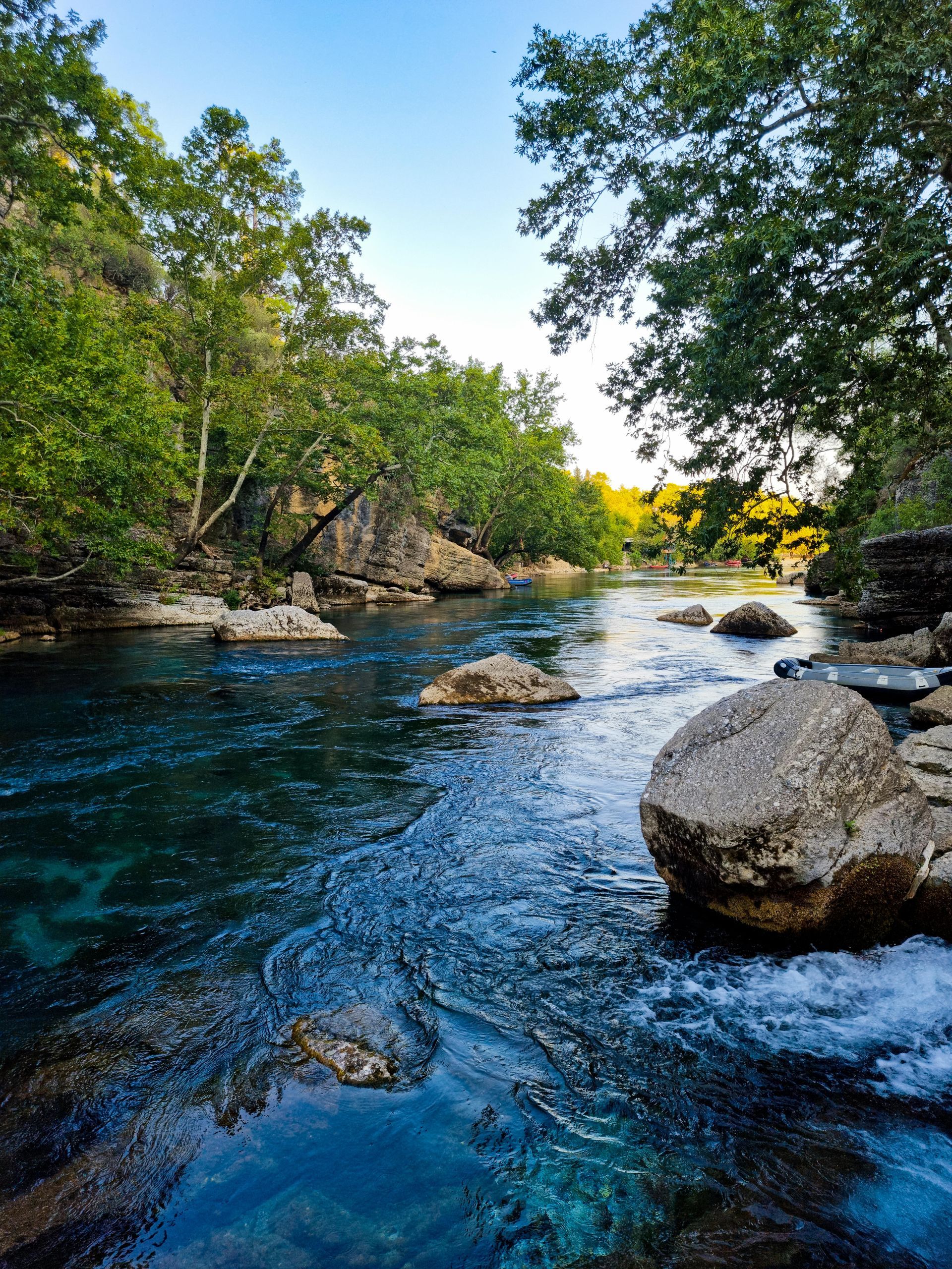 Rocky river with clear blue water, trees on both banks, and sunlight in the distance.