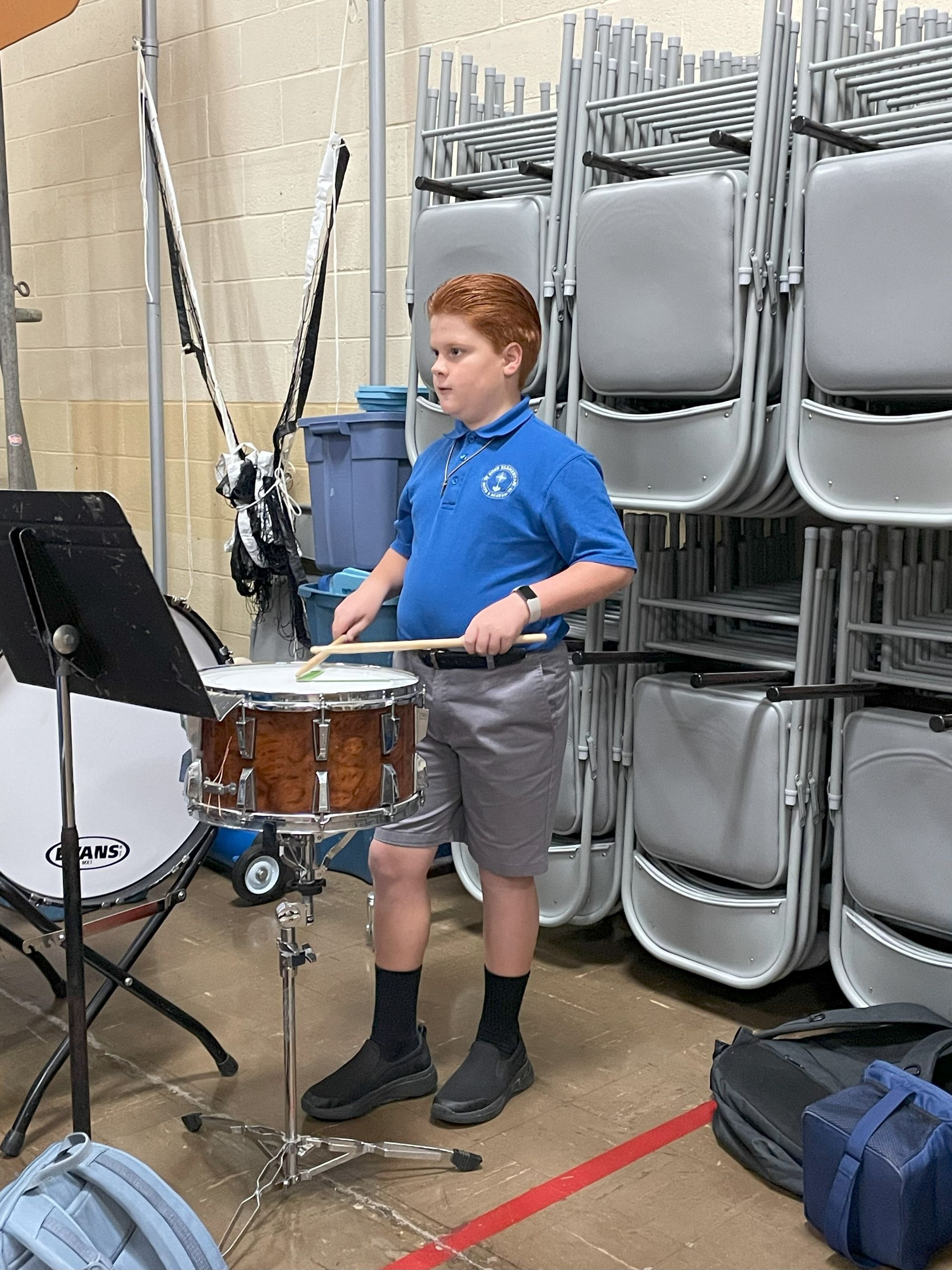 Boy with red hair playing a snare drum indoors. He wears a blue polo shirt, gray shorts, and black socks and shoes.