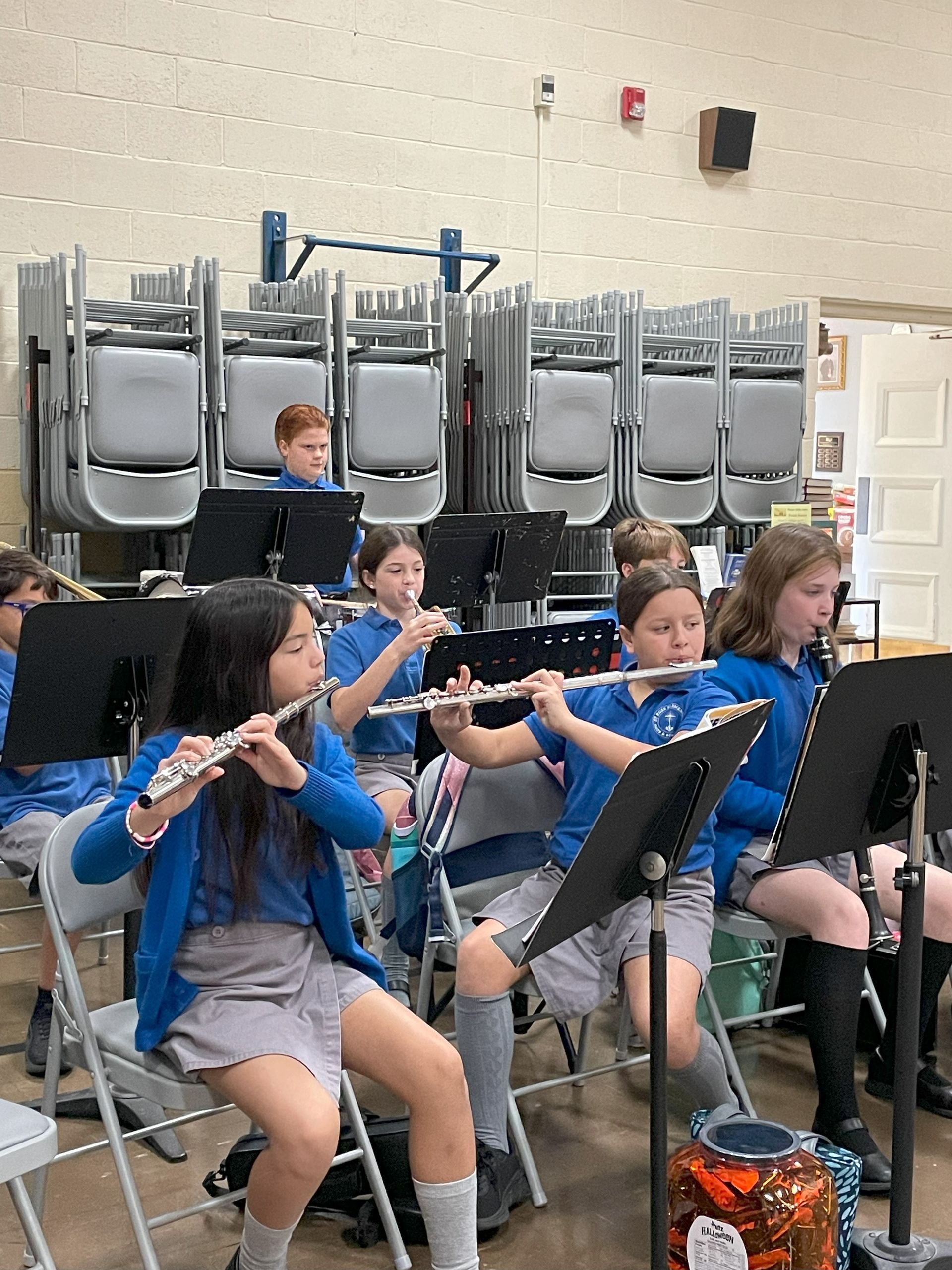 Children in a band, playing flutes. They are wearing blue shirts and gray skirts, sitting in chairs.