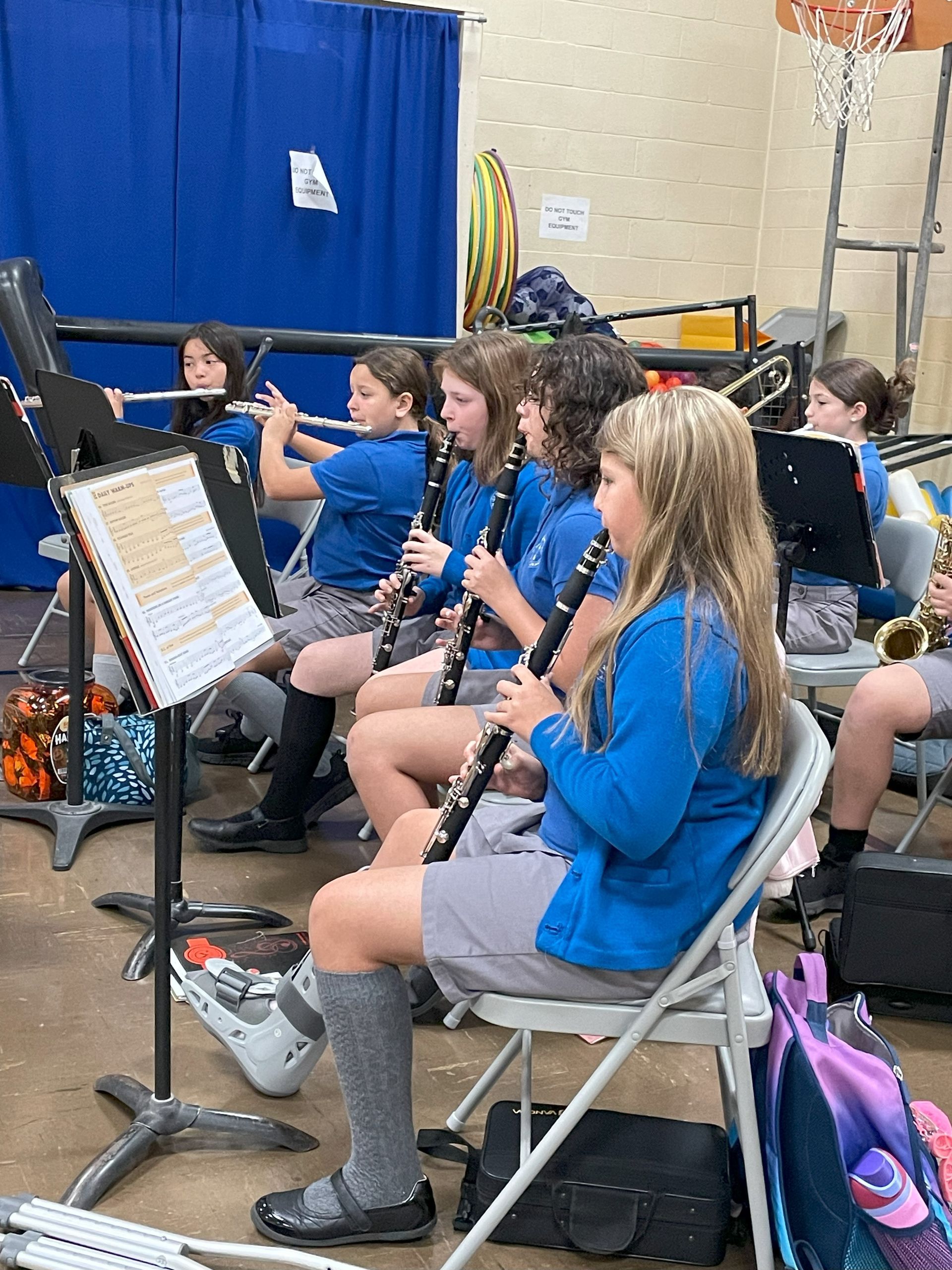 School band members play instruments, blue shirts, grey shorts, indoor setting.