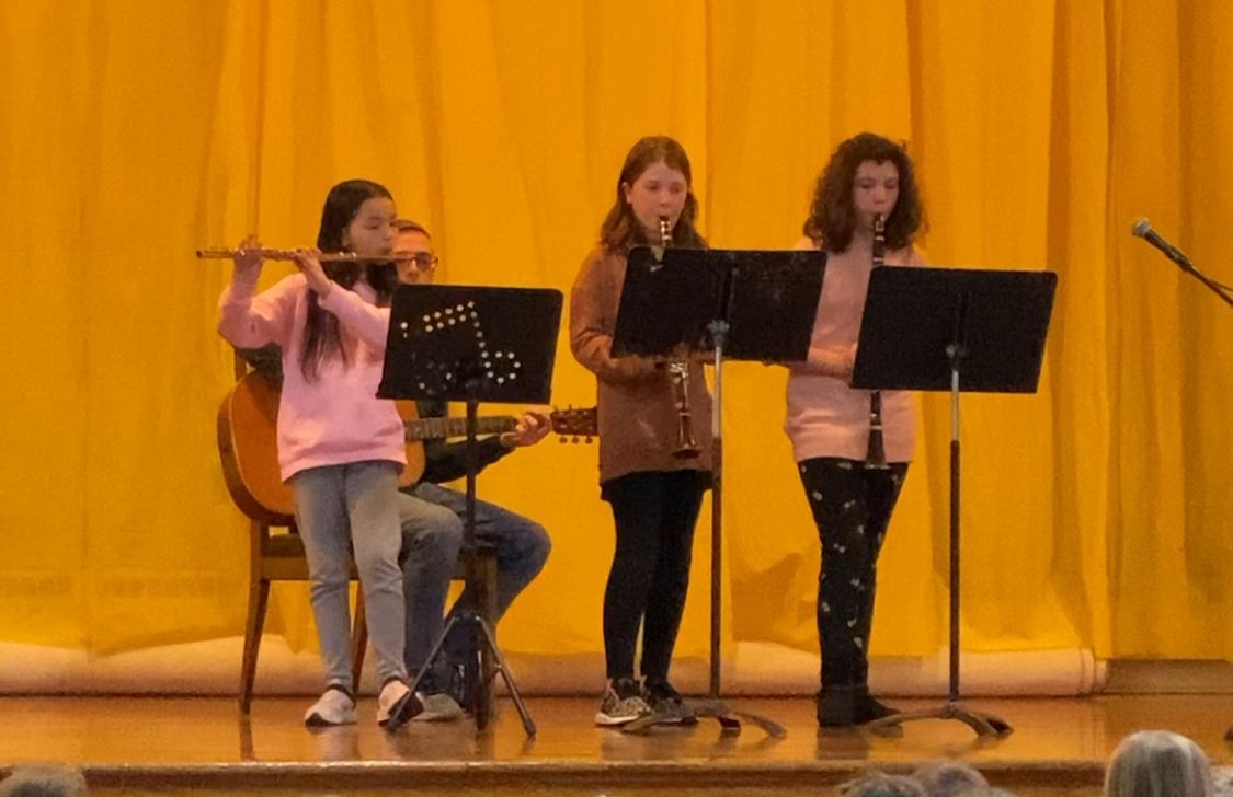 Musicians on stage: flute, clarinet, and guitar. Yellow backdrop.