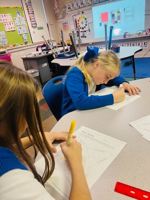 Two students in blue shirts drawing at a desk in a classroom; a color wheel is on the wall.