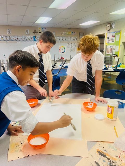 Three students in school uniforms work on a craft project at a table with paint and brushes.
