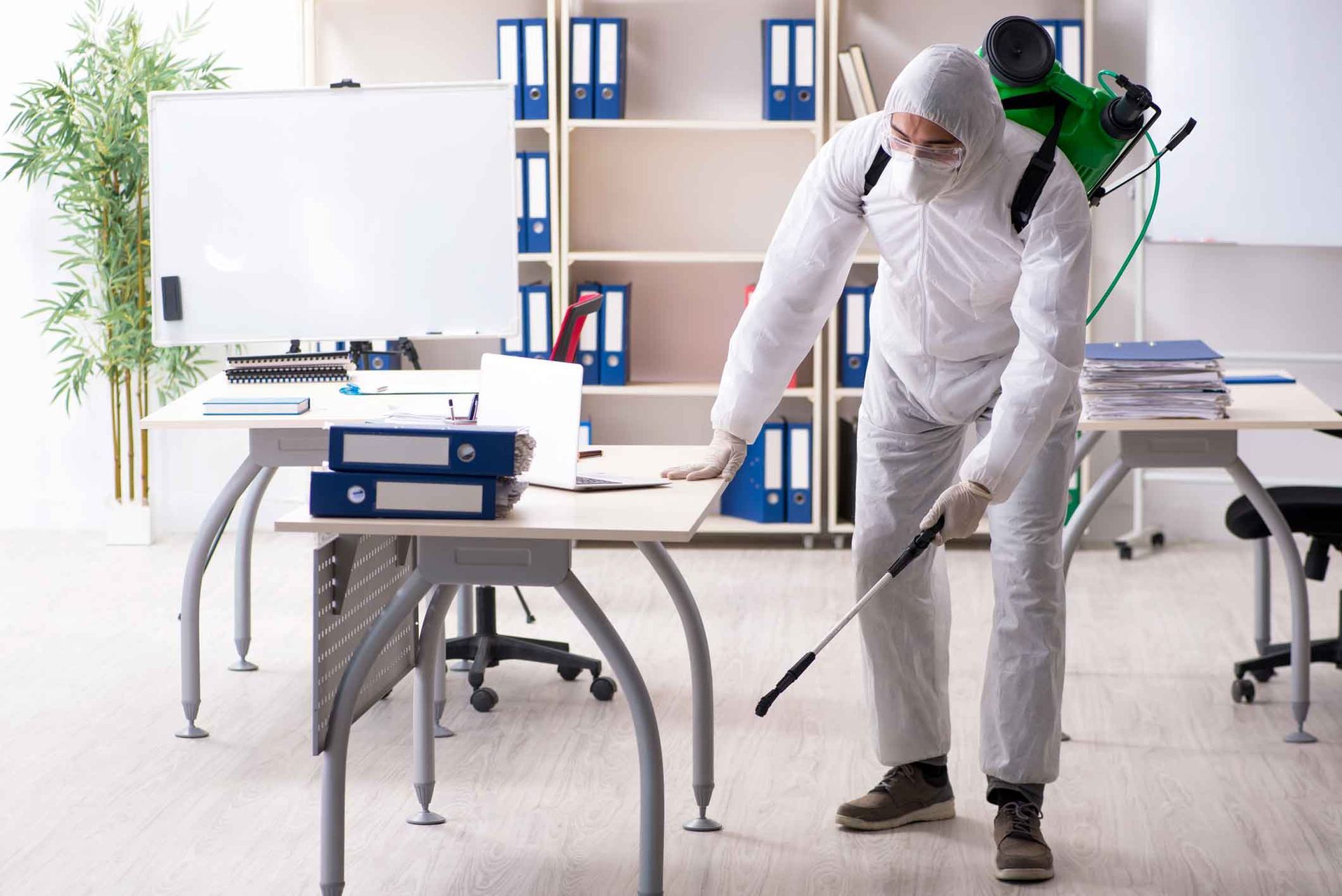 Worker in white protective suit disinfecting a bright office with a spray wand Worker in white protective suit disinfecting a bright office with a spray wand