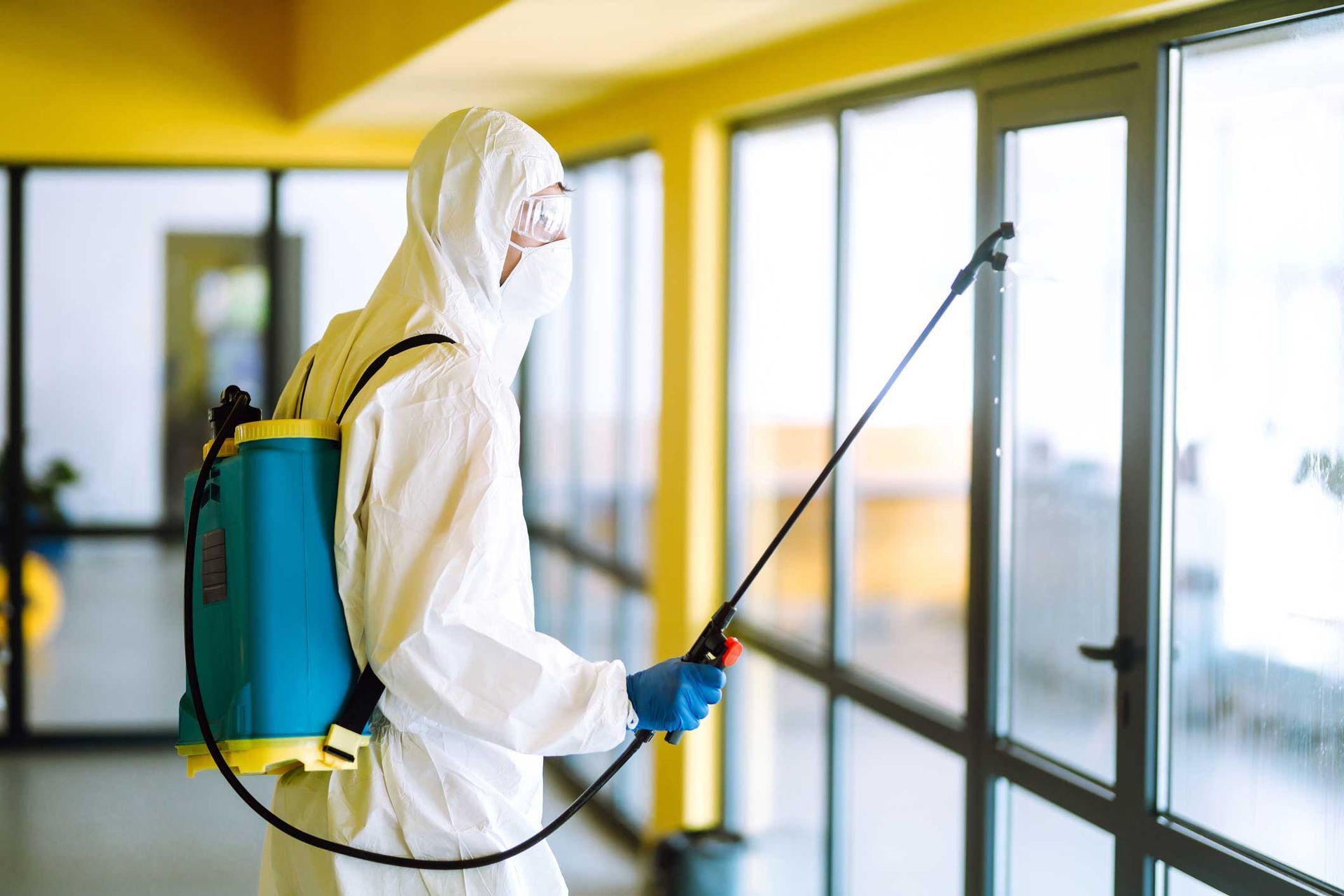Worker in protective suit spraying disinfectant on glass windows indoors