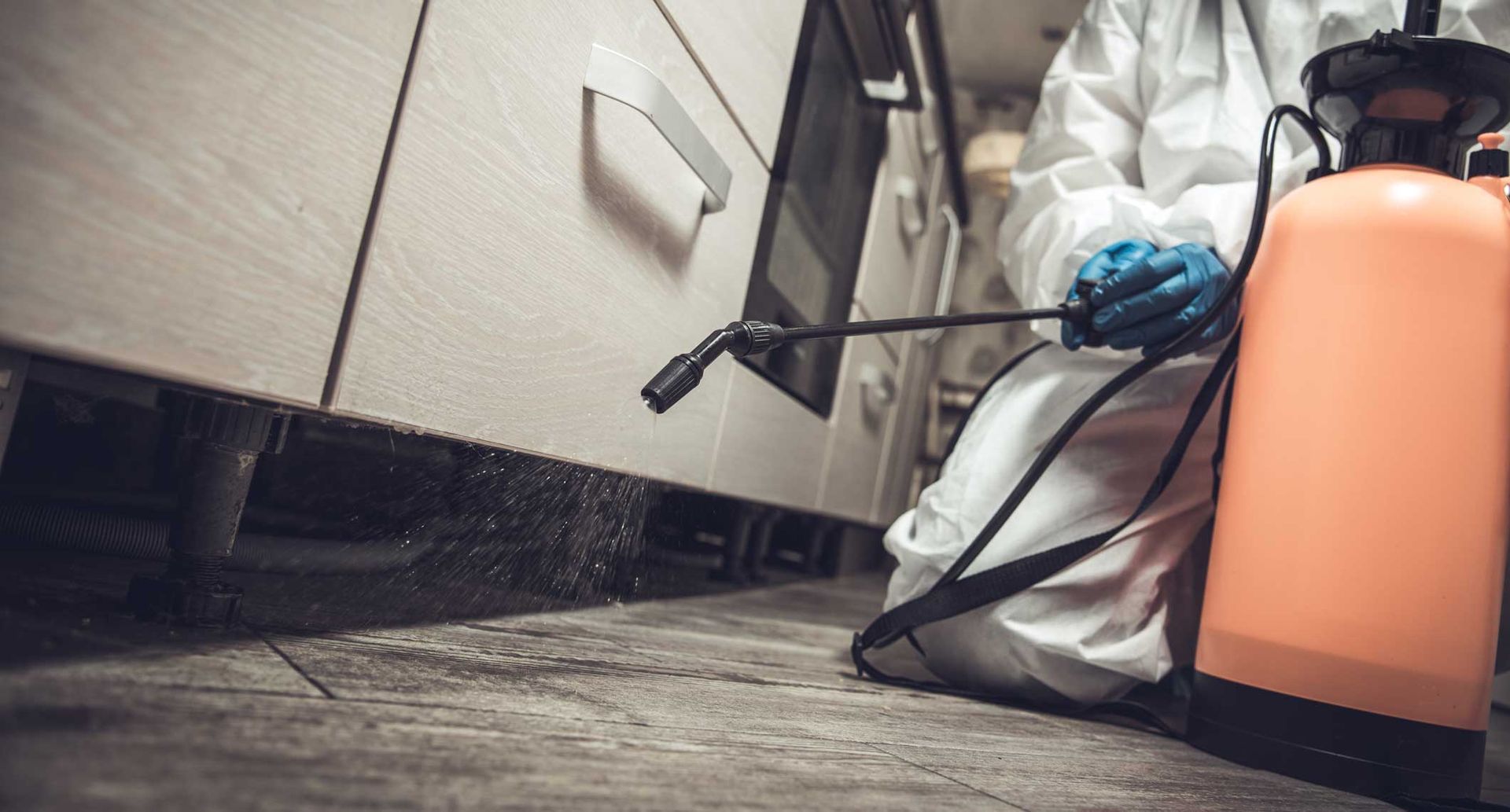 Worker in protective gear using a sprayer near a cabinet floor indoors