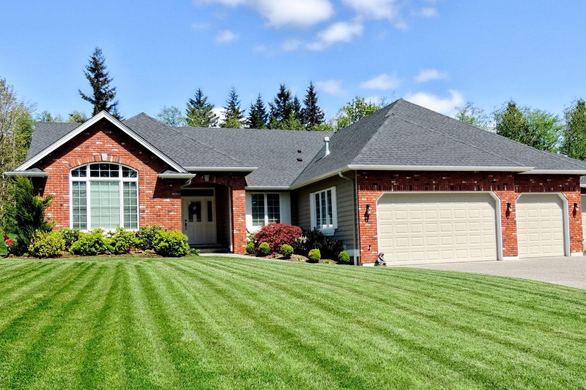 A single-story brick house with a two-car garage, a gray shingle roof, and a neatly mowed lawn under a clear blue sky.