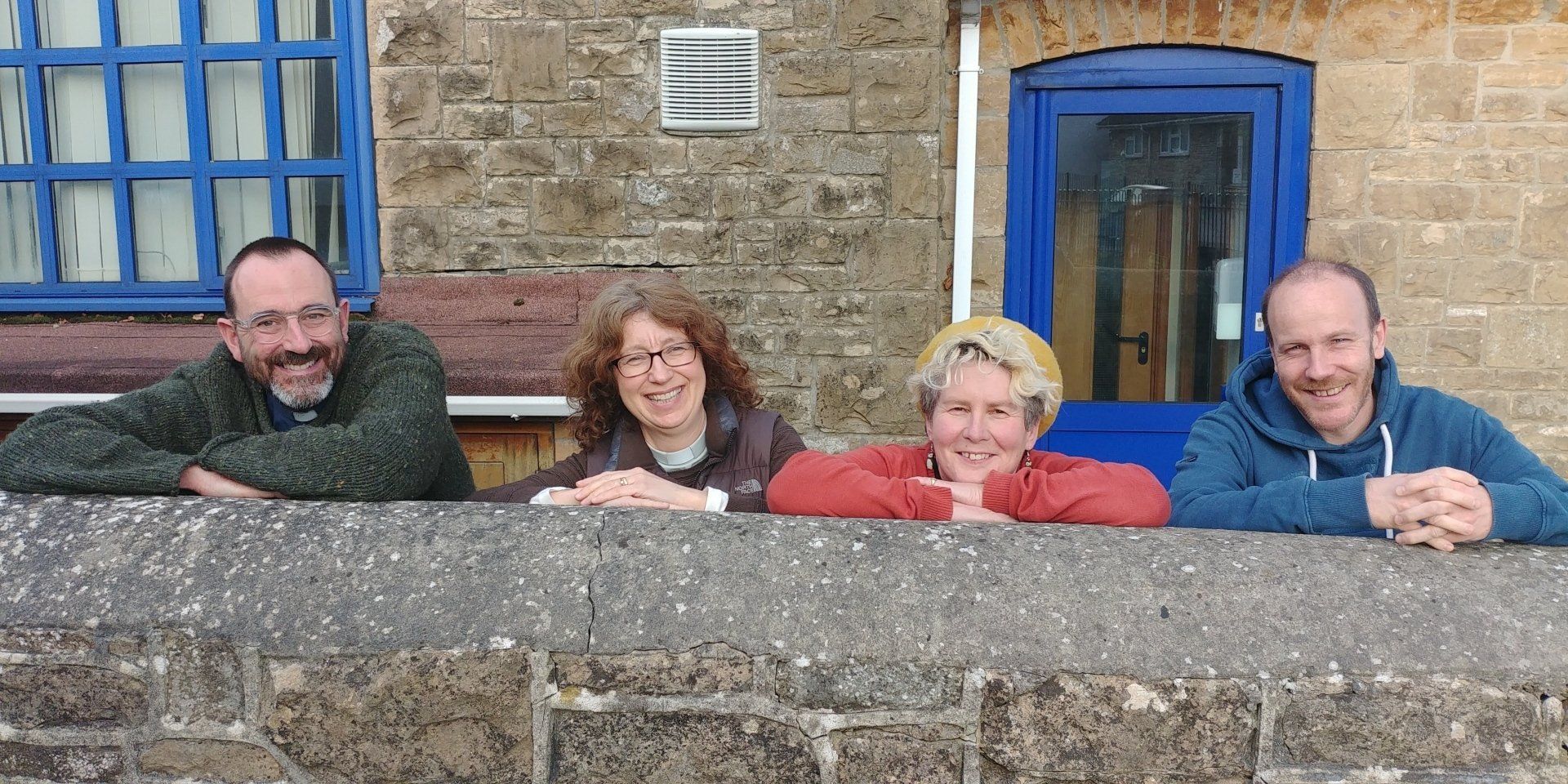 Four people leaning on a stone wall, smiling. Building with blue window and door in background.