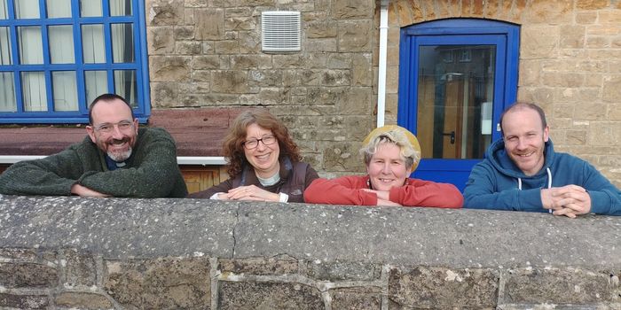 Four people leaning on a stone wall, smiling. Building with blue window and door in background.