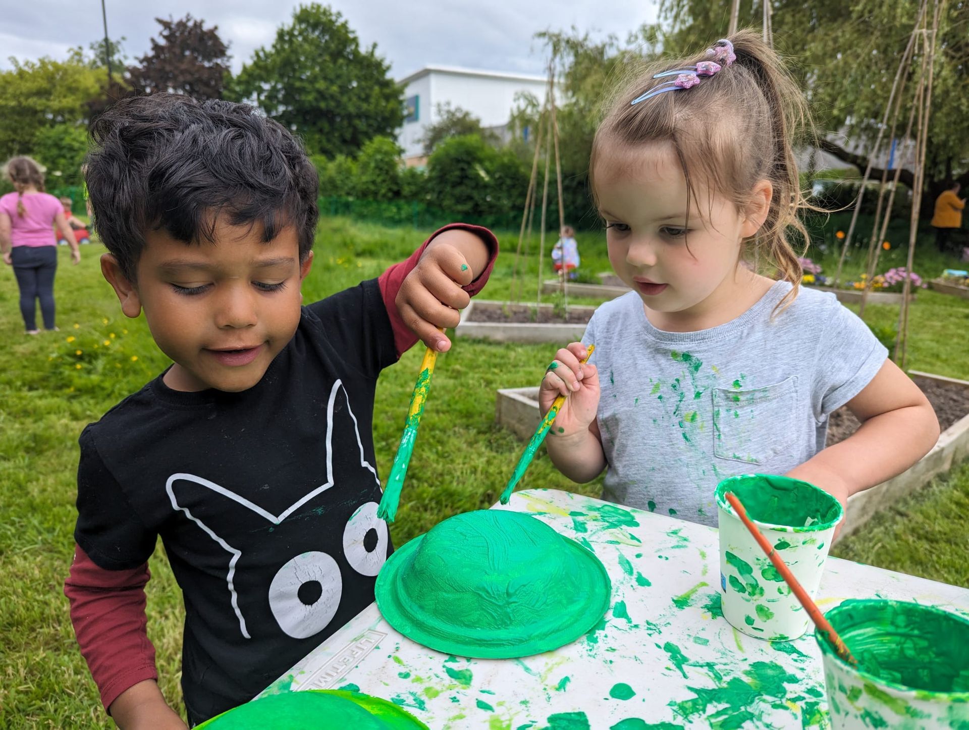 Two children painting green objects at a table outdoors.