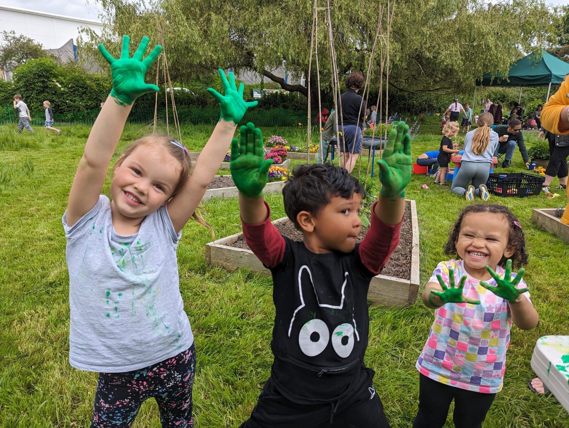 Three children in a garden, smiling, holding up hands covered in green paint. Other people and planters in background.