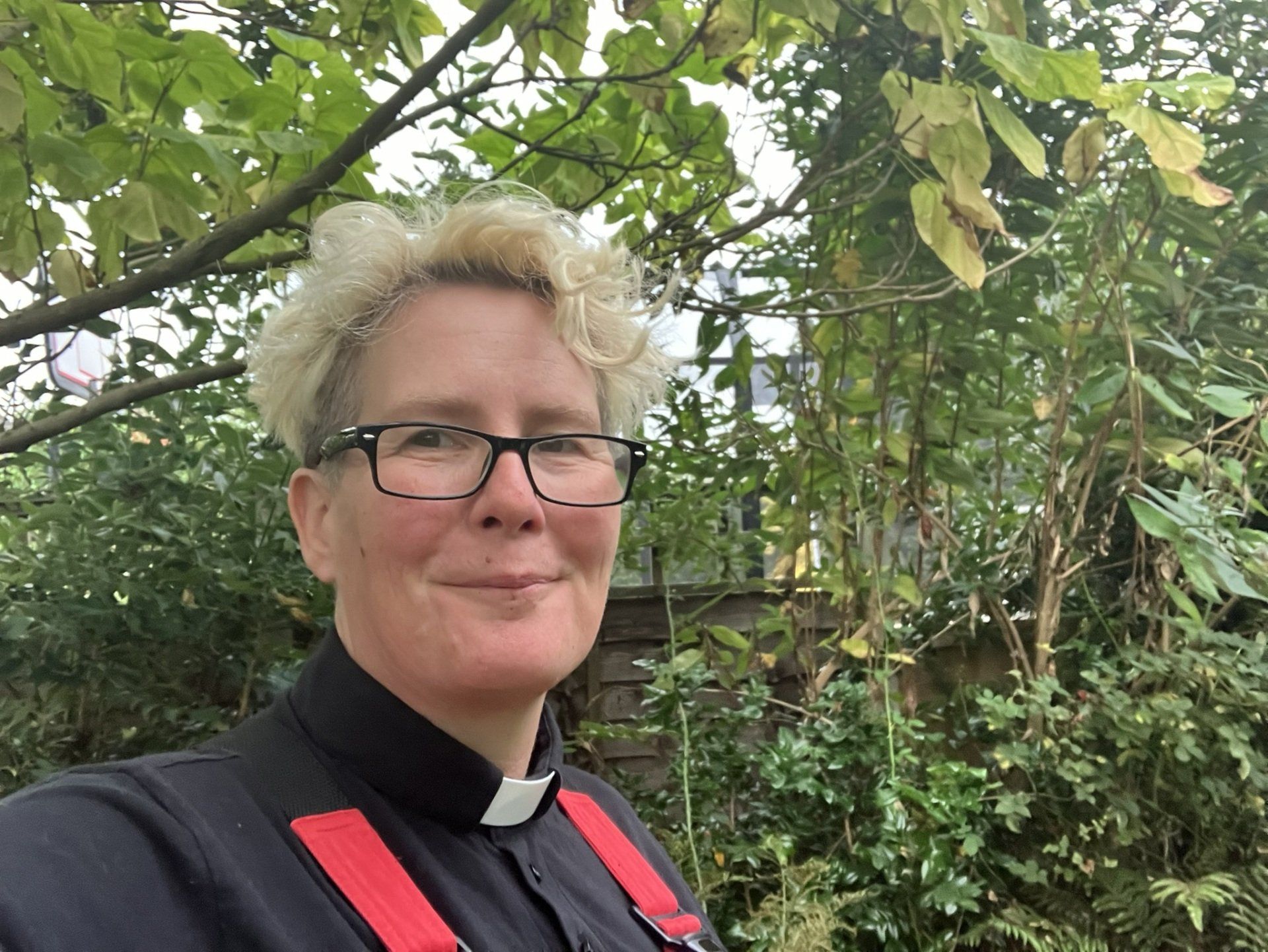 Person wearing glasses and clerical collar smiles in a garden. Red suspenders.